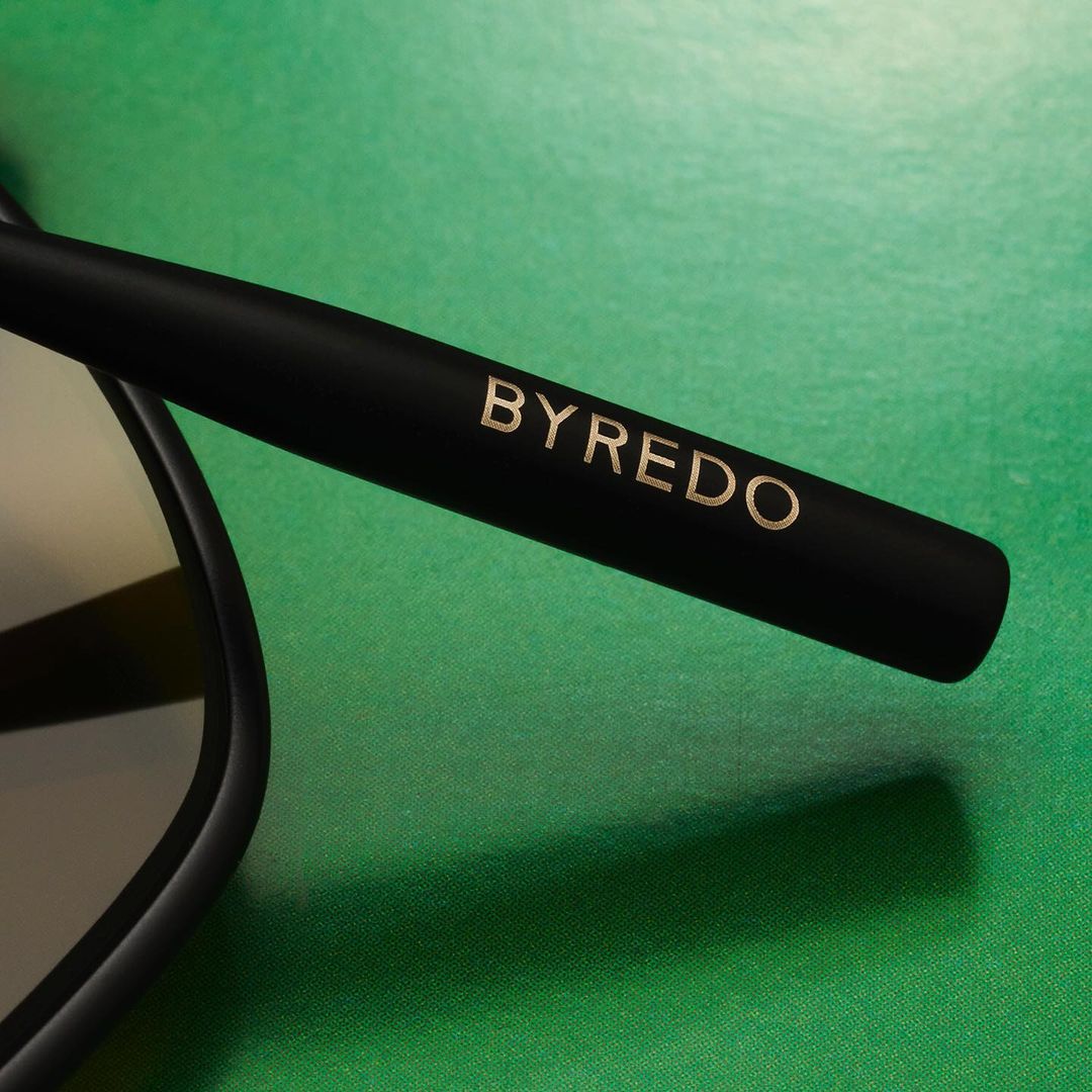 Close-up of black Byredo sunglasses arm with gold lettering on a textured green background in a product shot for fashion and retail.