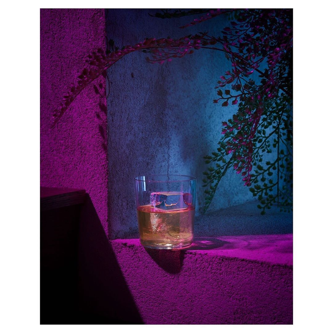 A rocks glass with amber liquid and a large ice cube, illuminated with purple and blue light, creating a vibrant, surreal aesthetic.