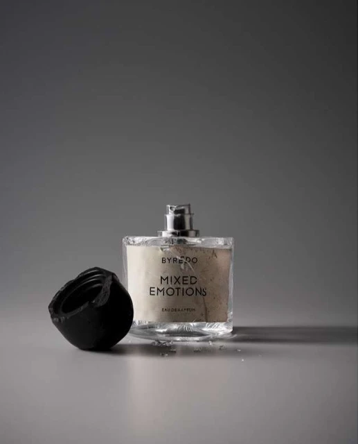 Byredo Mixed Emotions fragrance: clear glass bottle with black cap on gray surface. Modern beauty product shot. Cosmetic still life.