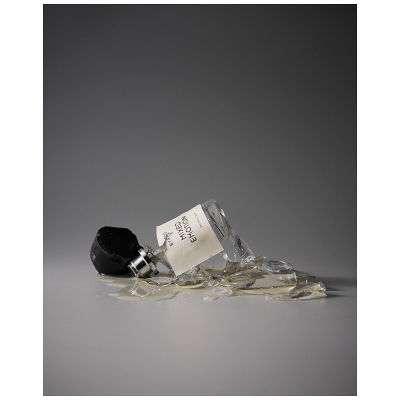 A shattered bottle of Byredo Mixed Emotions fragrance lies on its side, surrounded by broken glass on a gray surface and background.