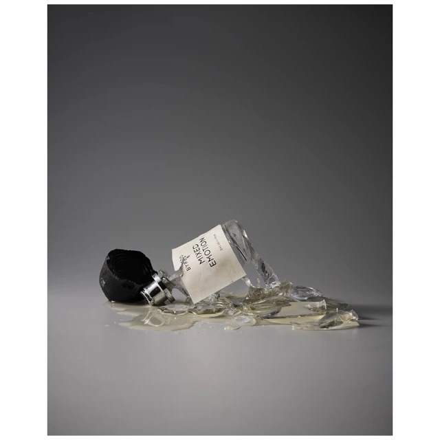 A shattered bottle of Byredo Mixed Emotions fragrance lies on its side, surrounded by broken glass on a gray surface and background.