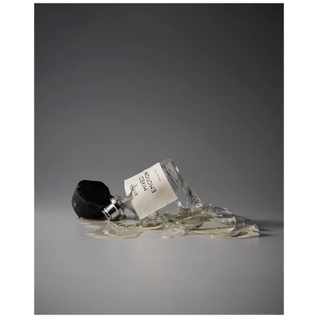 A shattered bottle of Byredo Mixed Emotions fragrance lies on its side, surrounded by broken glass on a gray surface and background.