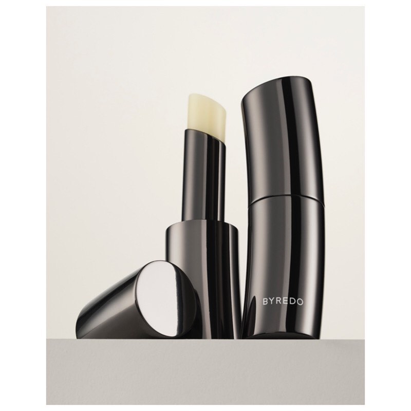 Close-up studio shot of Byredo lip balm. The sleek, gunmetal grey balm stick and container sit side by side on a neutral surface.