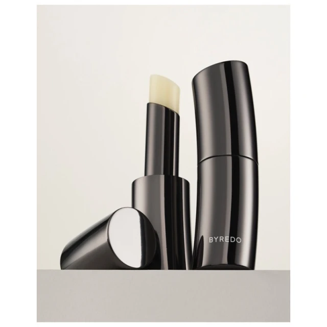 Close-up studio shot of Byredo lip balm. The sleek, gunmetal grey balm stick and container sit side by side on a neutral surface.