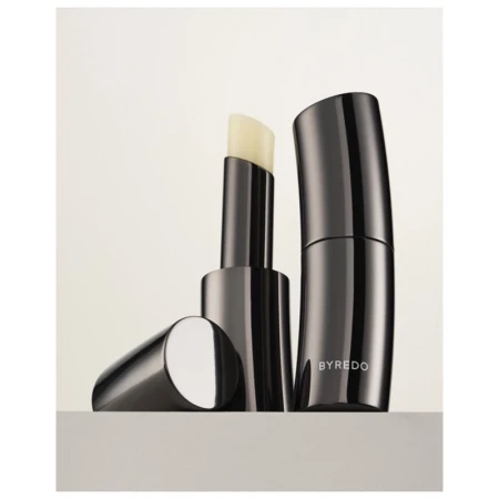 Close-up studio shot of Byredo lip balm. The sleek, gunmetal grey balm stick and container sit side by side on a neutral surface.
