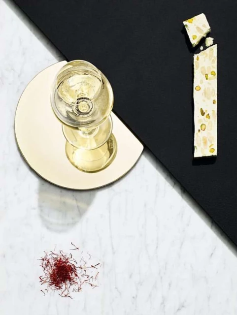Overhead shot of a clear wine glass on a gold coaster, white nougat candy, and saffron threads against a split marble and black background.