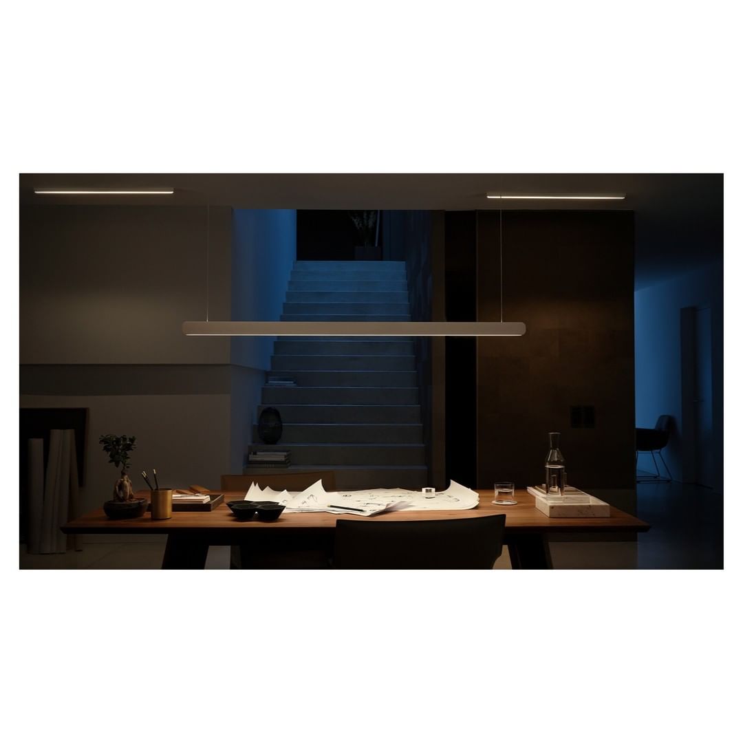 Illuminated linear pendant lighting fixture design hanging over a modern dining table inside a room with a staircase in the background.