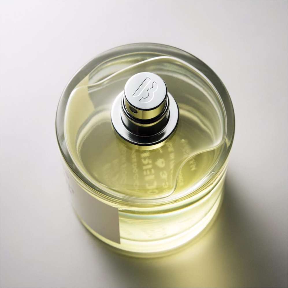 Overhead studio shot of a transparent glass perfume bottle with a silver cap, liquid inside, and blank white label on a gradient background.