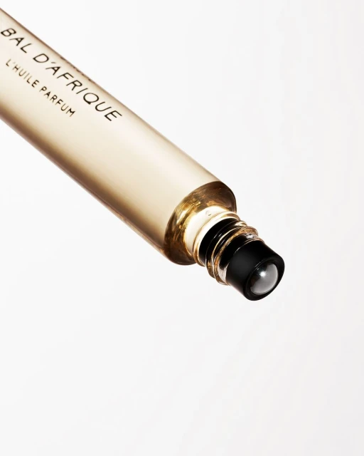 Close-up of a clear, slender roll-on perfume bottle with gold accents and black cap, featuring the label 'Bal D'Afrique L'Huile Parfum'.
