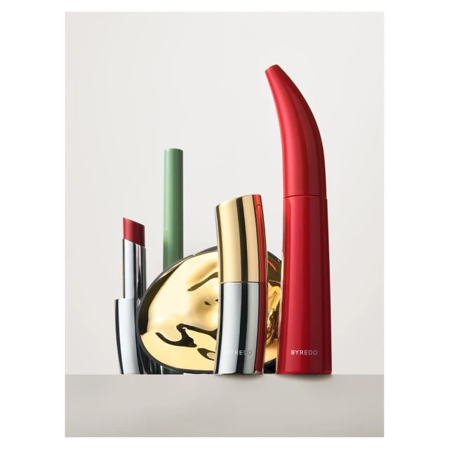 Close-up product shot of Byredo lipsticks, lip balm, and mirror compact in red, gold, green, and silver against a gradient background.