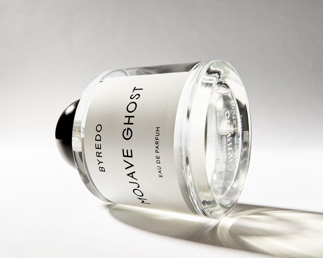 A glass bottle of Byredo Mojave Ghost perfume is laying on its side against a white background with a soft shadow.