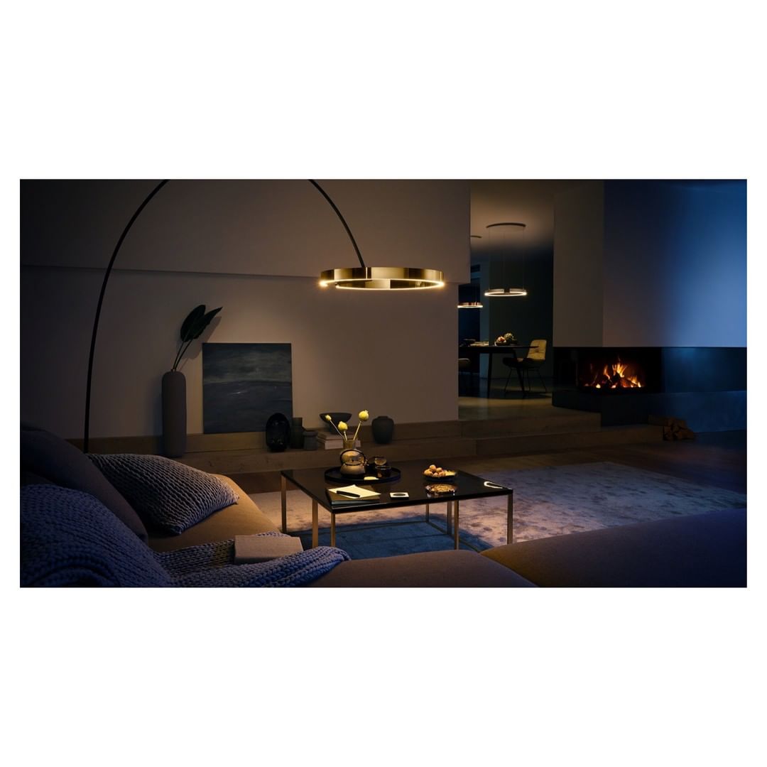 Interior with modern gold ring lamp over a brown sofa, coffee table with flowers, and fireplace. Warm lighting and soft textures.