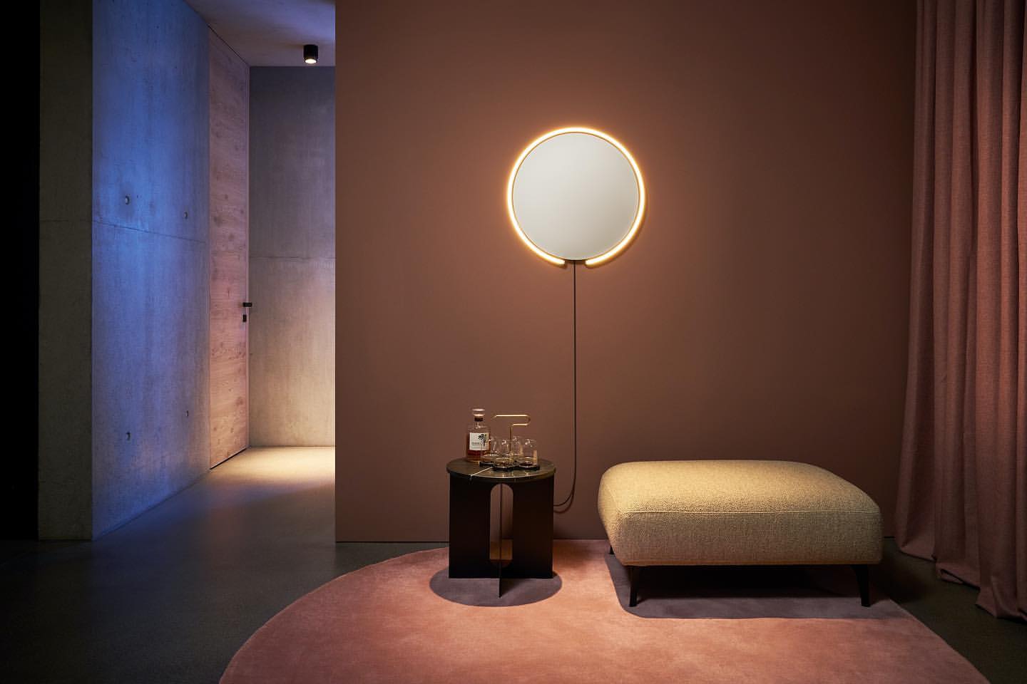 Interior design featuring a round illuminated mirror above a small table with glassware, with an ottoman on a pink round rug.