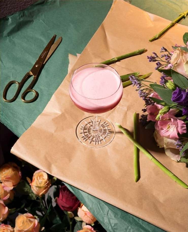 Overhead shot of a pink cocktail on craft paper, surrounded by flowers, green stems, and gold scissors against a teal background.