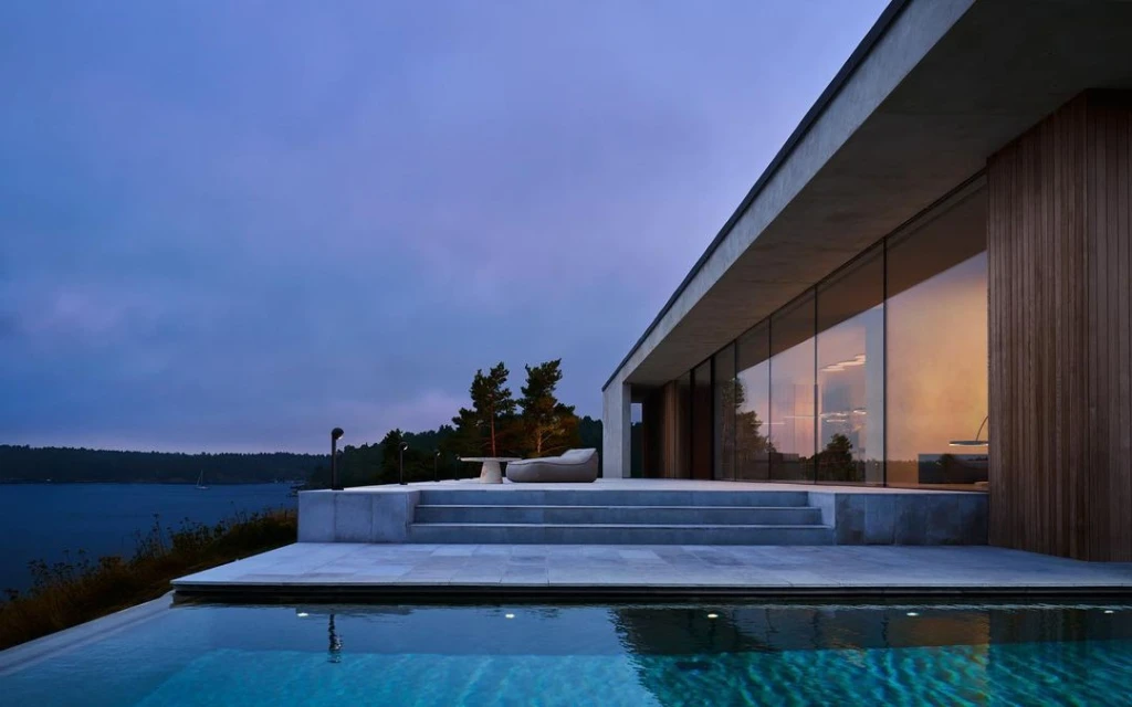 Exterior view of modern house with infinity pool, concrete patio and lounge furniture against a backdrop of a forest and dusk sky.