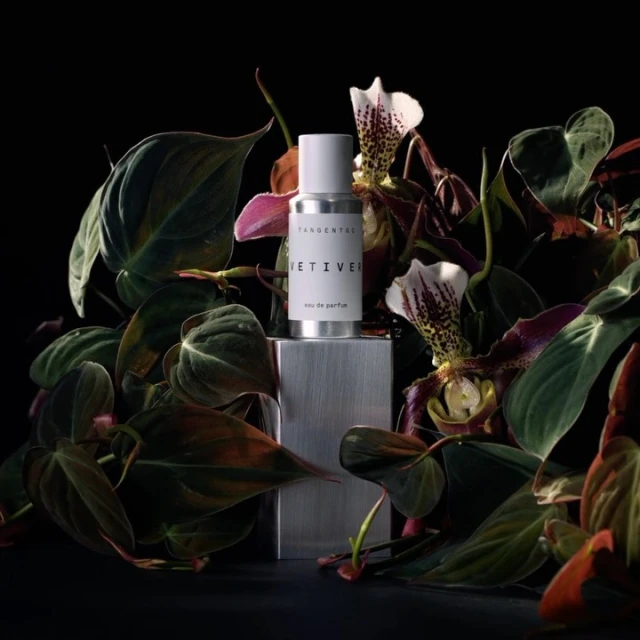 Perfume bottle by Tangent GC, 'Vetiver' scent, centered on a metallic block surrounded by green foliage and exotic orchid flowers, studio lit.