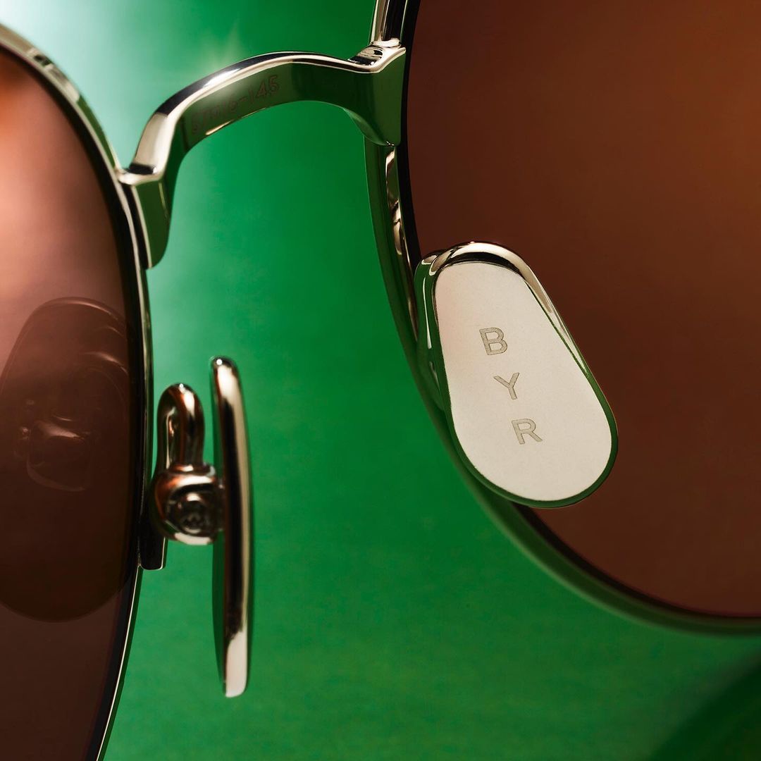 Close-up of sunglasses with a gold frame and brown lenses against a green background with the text 'BYR' visible on the nose pad.