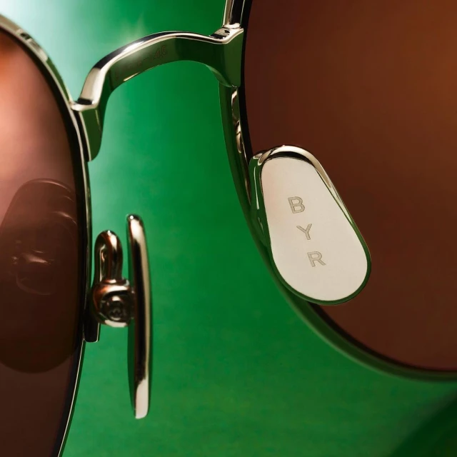 Close-up of sunglasses with a gold frame and brown lenses against a green background with the text 'BYR' visible on the nose pad.