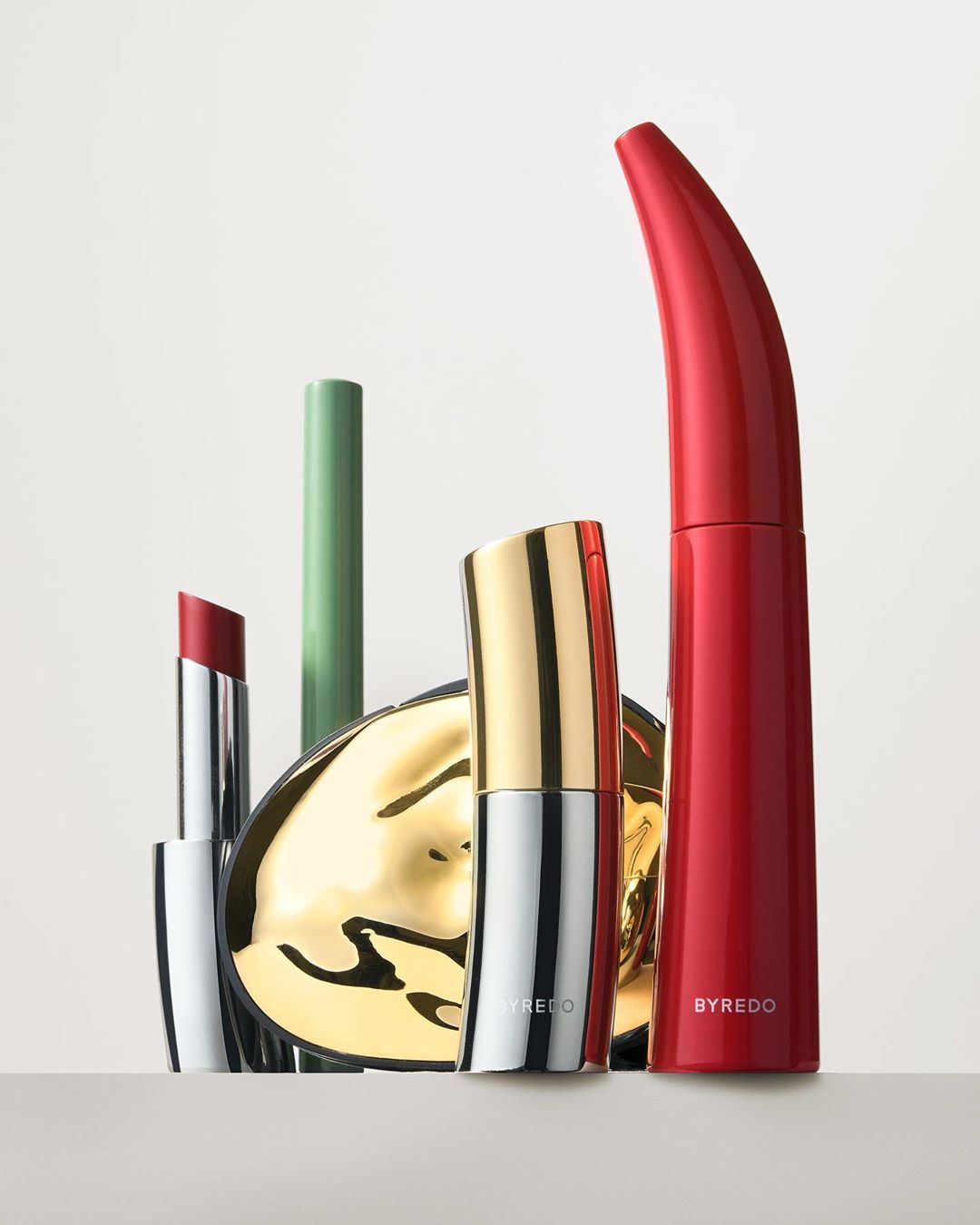 Byredo makeup collection featuring lipstick, mascara, and a compact in silver, gold, red, and green displayed in a light studio setting.