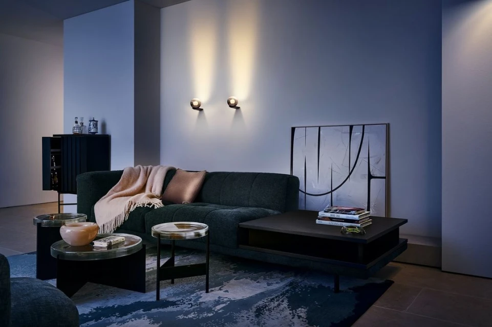 Living room interior features a green sofa with a throw and pillow, flanked by side tables, a rug, and wall-mounted lighting fixtures.