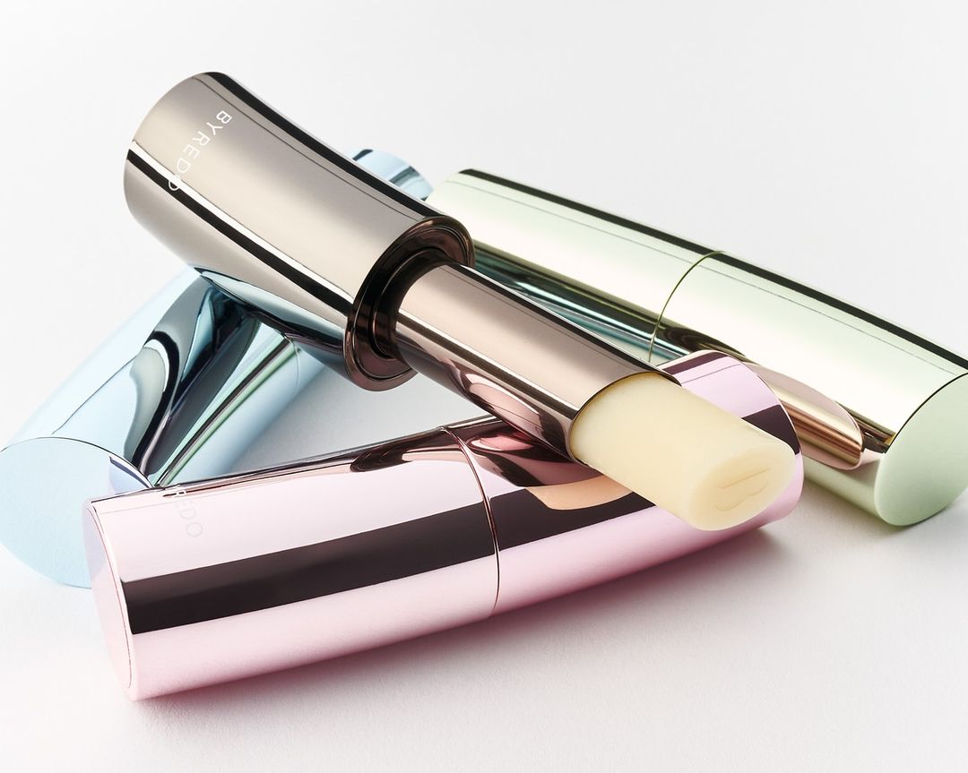 Close-up of three pastel-colored Byredo lip balms arranged on a white surface with studio lighting, showcasing their sleek, metallic packaging.