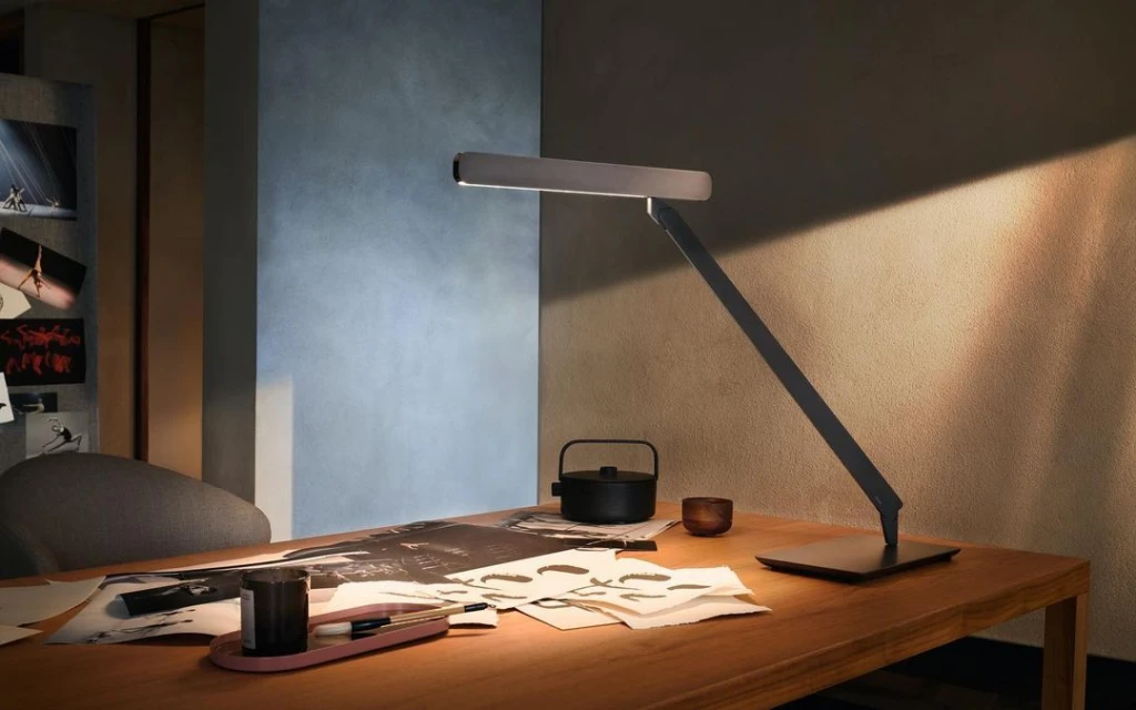 A modern adjustable task lamp illuminates a wooden desk with sketches and art supplies indoors, casting a warm glow.