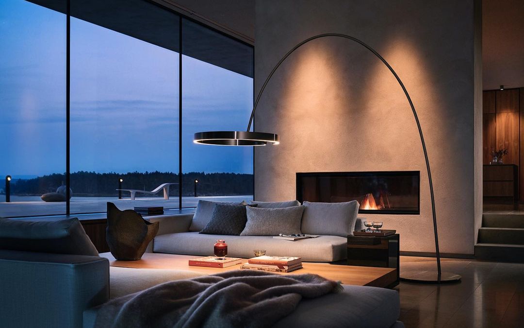Modern living room interior with a gray sofa, contemporary lighting, fireplace, and floor-to-ceiling windows at dusk. Interior design.