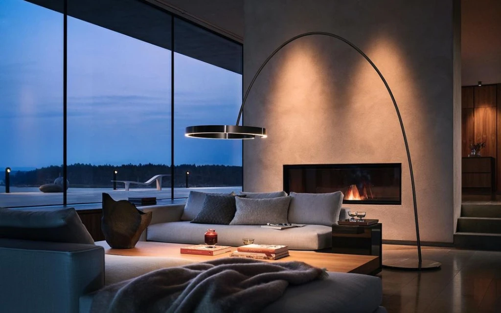 Modern living room interior with a gray sofa, contemporary lighting, fireplace, and floor-to-ceiling windows at dusk. Interior design.