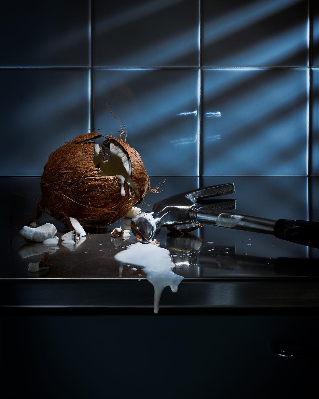 Still life shot of a cracked coconut and hammer on a dark countertop with a tiled backsplash, illuminated by light stripes.