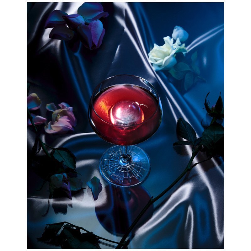 Overhead shot of a glass of red liquid with a sphere of ice and scattered flowers, set against a draped satin background.