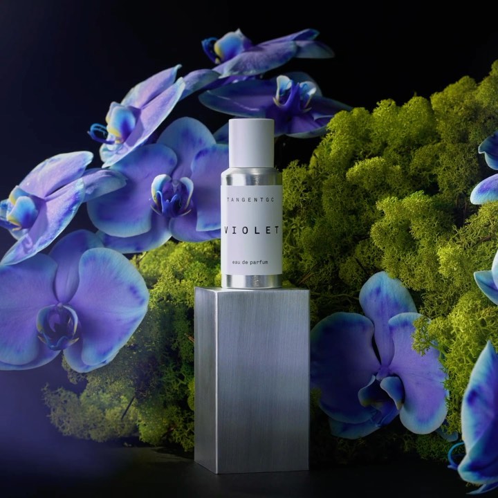 Elegant presentation of a silver perfume bottle on a metal stand, accented by blue orchids and vibrant green moss against a black backdrop.
