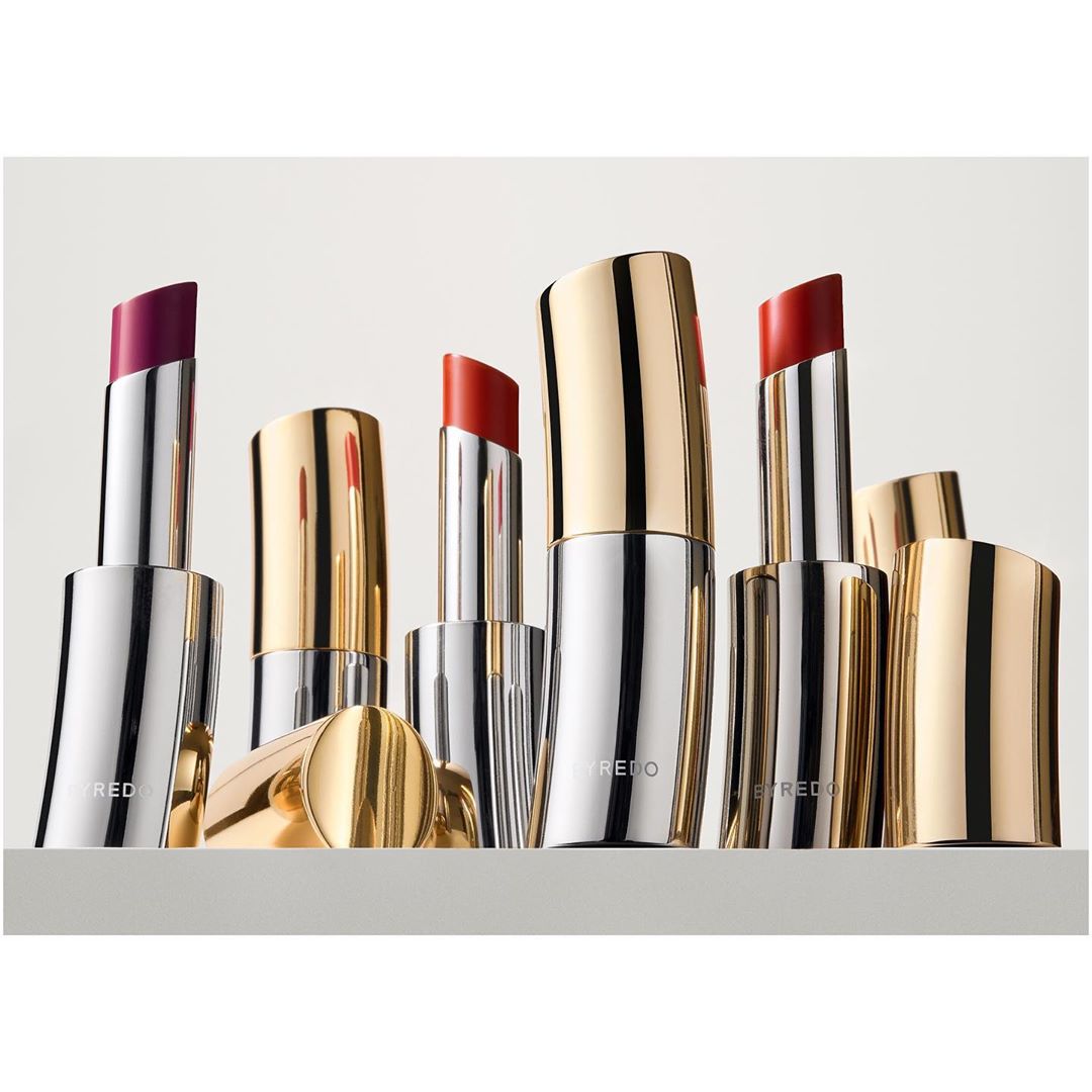 Assorted lipsticks with silver and gold casings are arranged on a gray surface against a bright background, offering a variety of colors.