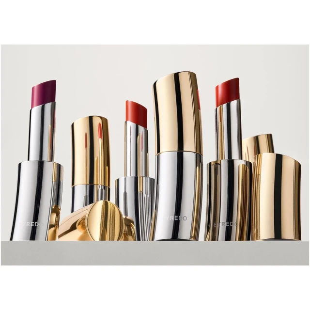 Assorted lipsticks with silver and gold casings are arranged on a gray surface against a bright background, offering a variety of colors.