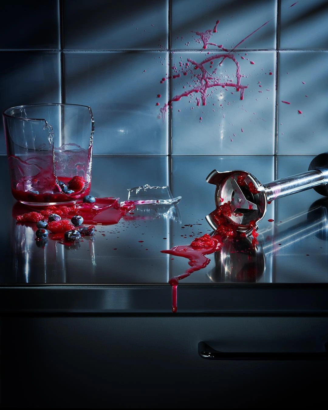 Broken glass blender jar and immersion blender surrounded by spilled red berry smoothie on a metallic kitchen counter.