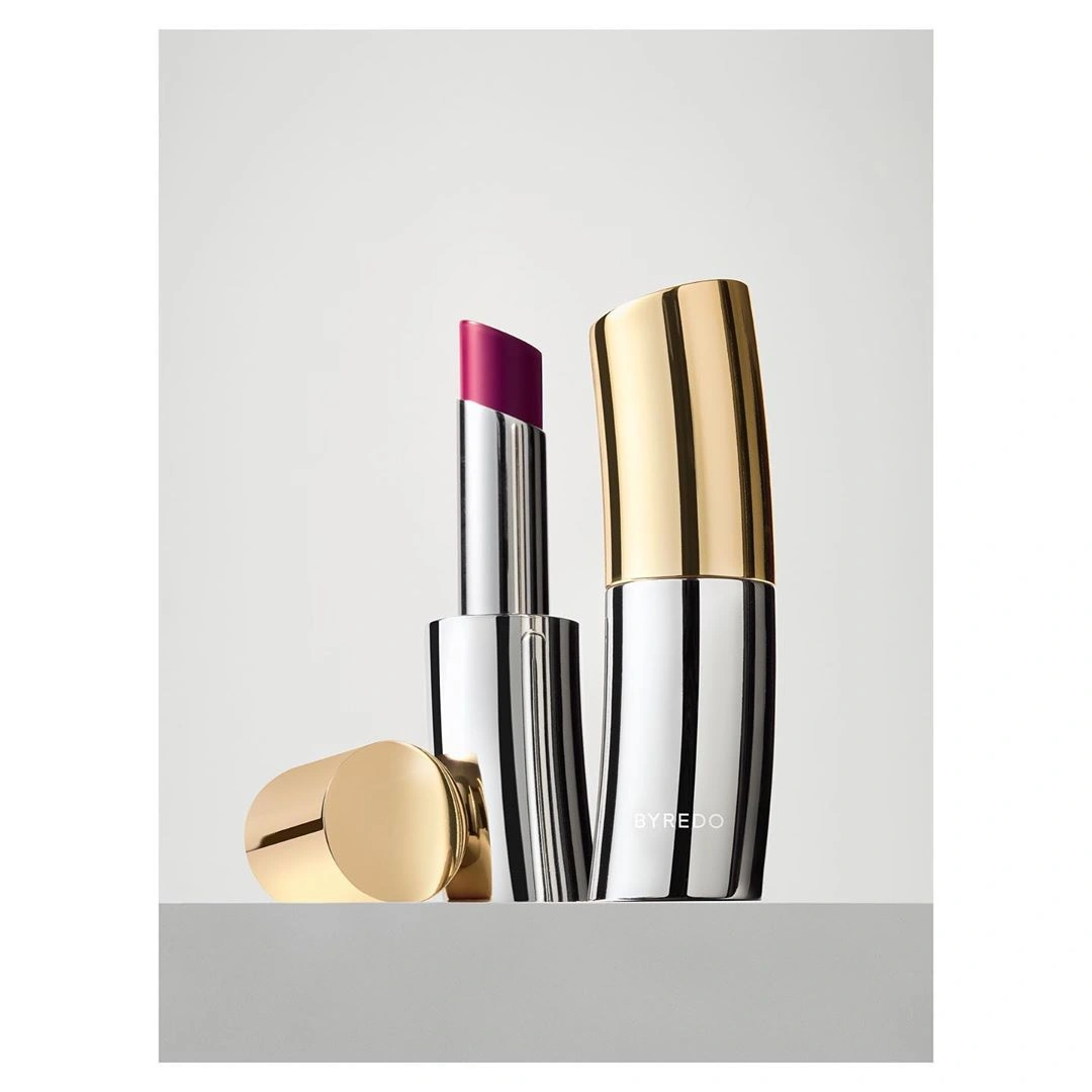 Byredo lipstick in a curved metallic silver and gold case with a bright magenta bullet against grey