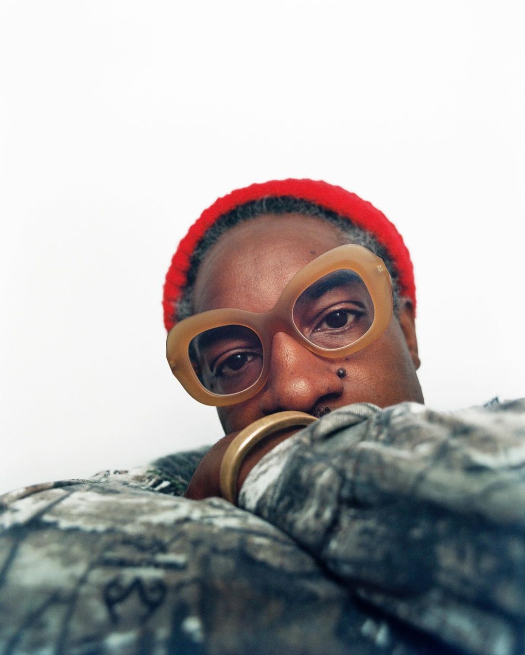 Close-up portrait of an adult wearing beige-framed glasses and a red beanie, gazing directly at the camera with a neutral expression.