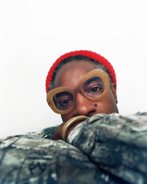 Close-up portrait of an adult wearing beige-framed glasses and a red beanie, gazing directly at the camera with a neutral expression.