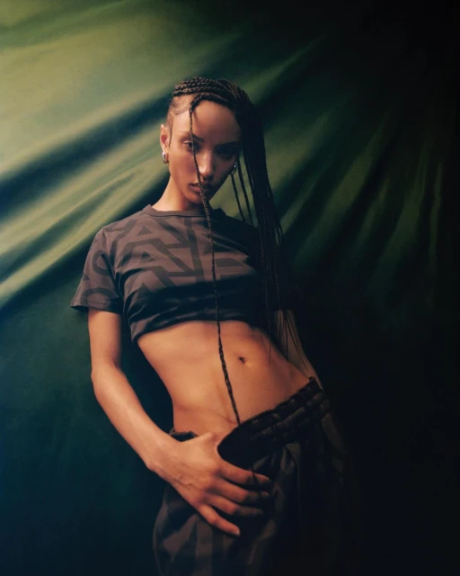 A young woman poses in a crop top and sweats against a draped green backdrop. Braided hair and a sultry facial expression.