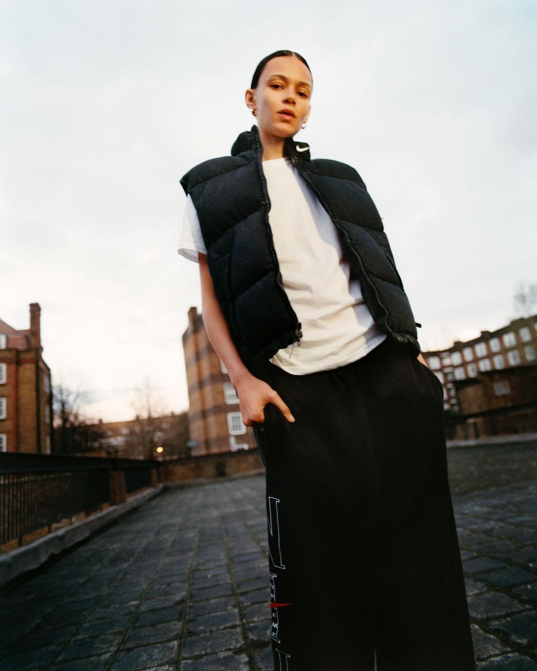 Woman poses on city rooftop wearing white t-shirt and black puffer vest. Buildings and sky in background, low-angle view, medium shot.
