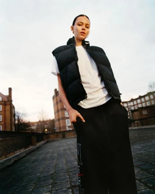 Woman poses on city rooftop wearing white t-shirt and black puffer vest. Buildings and sky in background, low-angle view, medium shot.