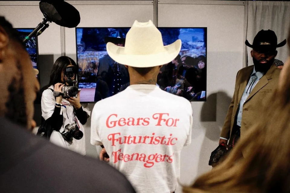 Fashion show backstage featuring a person in a cowboy hat, camerawoman with professional equipment, and a T-shirt with retro text.