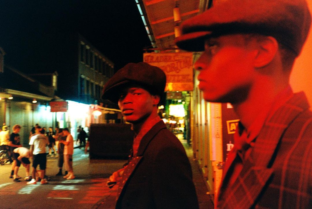 Two stylish men in suits and caps stand on a city street at night, illuminated by the vibrant lights of the nightlife.