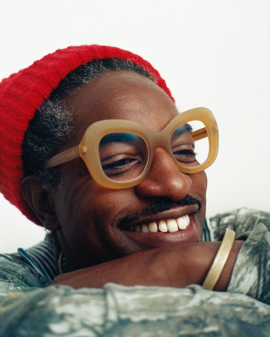 Close-up portrait of an adult male with mustache, wearing red knit cap and glasses, smiling slightly, with a patterned shirt and gold bracelet.