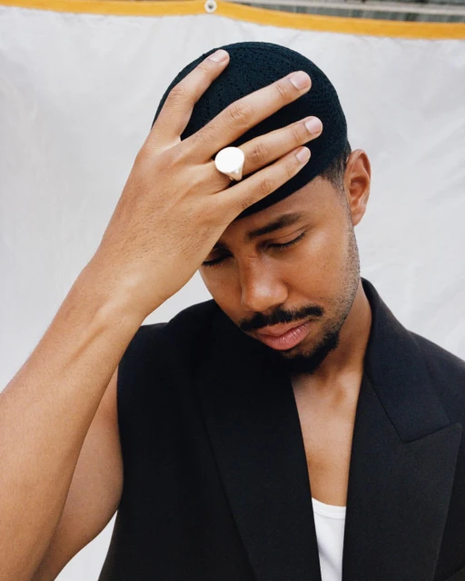 Portrait of a Black man wearing a black cap and vest with a white ring, hand touching his forehead, neutral expression, against a white backdrop.