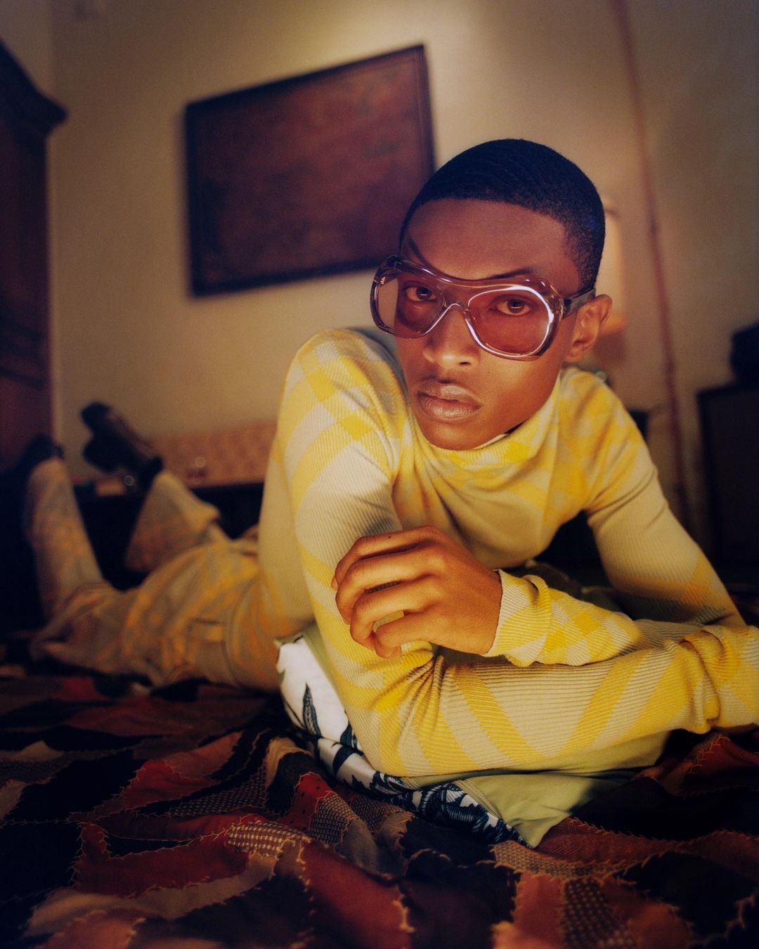 Person in yellow turtleneck and pants with sunglasses lying on a patterned bedspread with a pillow and a framed painting behind them.