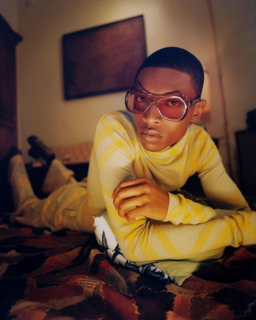 Person in yellow turtleneck and pants with sunglasses lying on a patterned bedspread with a pillow and a framed painting behind them.