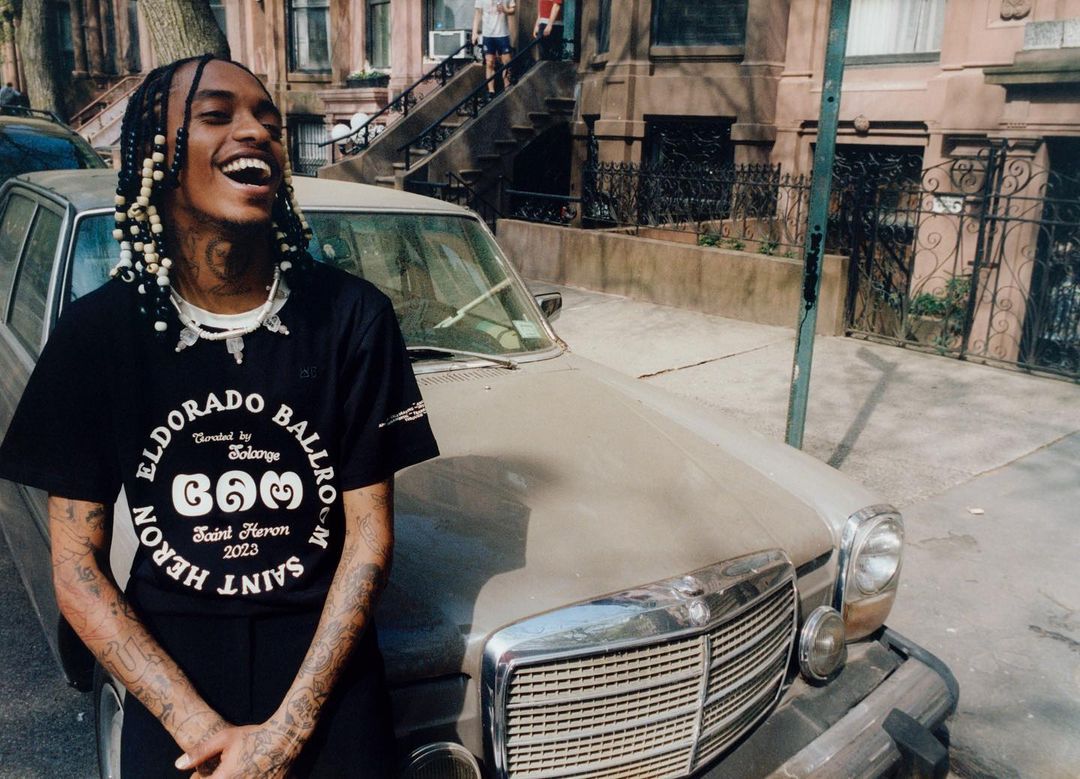 Adult male smiles next to a vintage car on a city street. The man wears a branded black t-shirt and beaded braids in an outdoor setting.