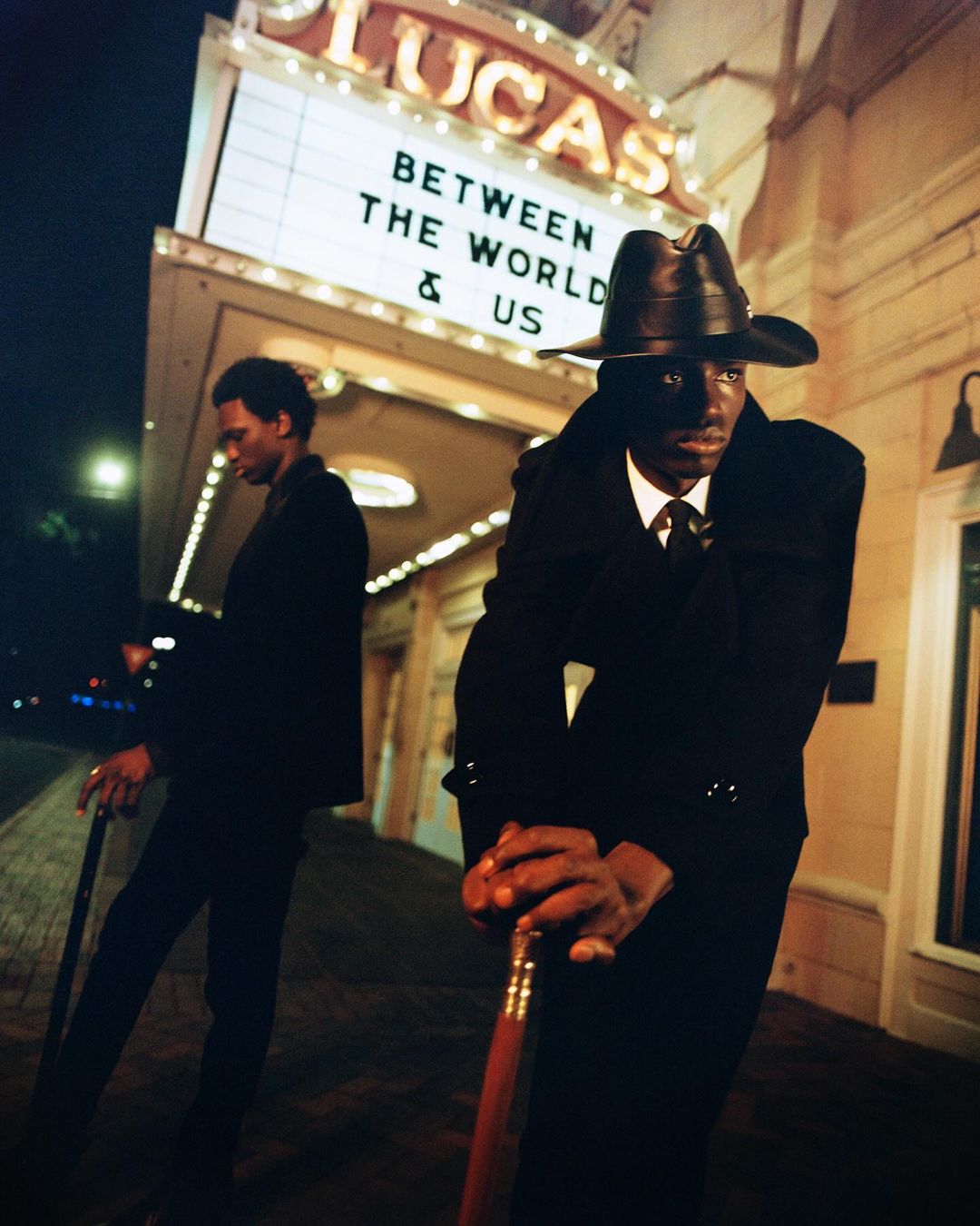 Two African-American men in dark suits and hats stand outside the Lucas Theatre at night, holding canes, with marquee lights illuminating them.