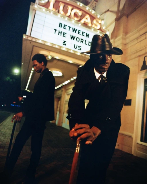 Two African-American men in dark suits and hats stand outside the Lucas Theatre at night, holding canes, with marquee lights illuminating them.