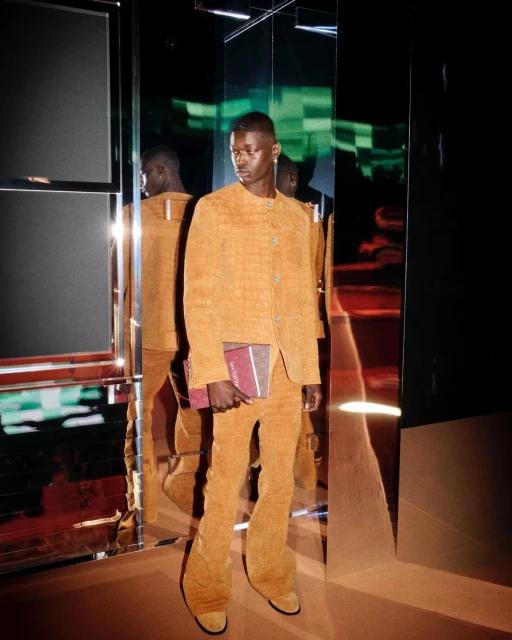 A Black male model poses indoors in a yellow corduroy suit, holding a burgundy book, reflecting in mirrored panels. The lighting is studio.